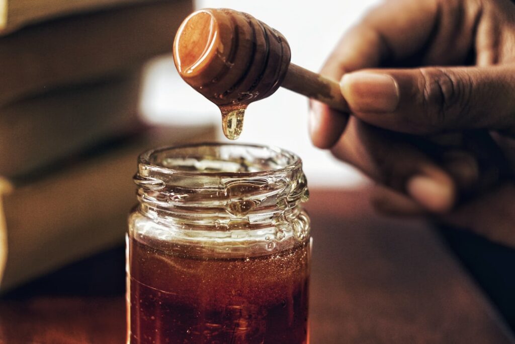A jar of golden natural honey with a wooden dipper resting on its rim surrounded by a warm inviting glow