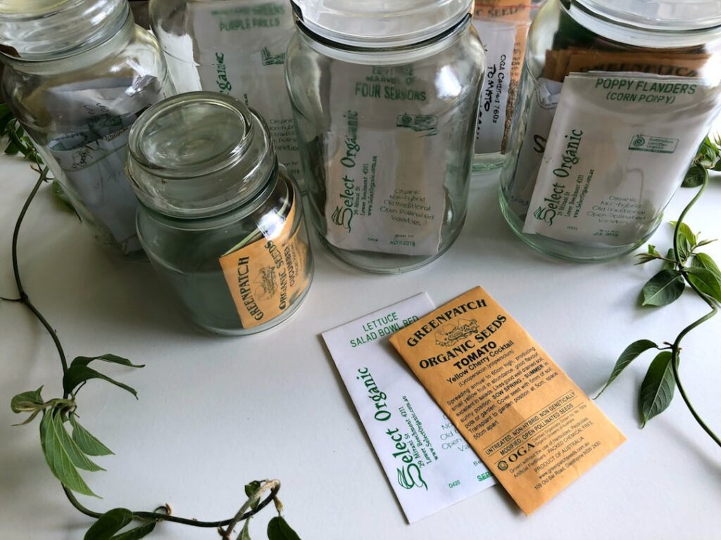 A collection of various dried vegetable and flower seeds in small glass jars or envelopes labeled with their names and d