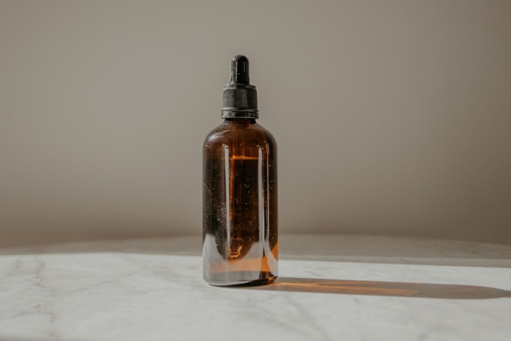 A glass jar of rice water next to a small bottle of baby oil infused with cloves with a spoon nearby on a dark counterto