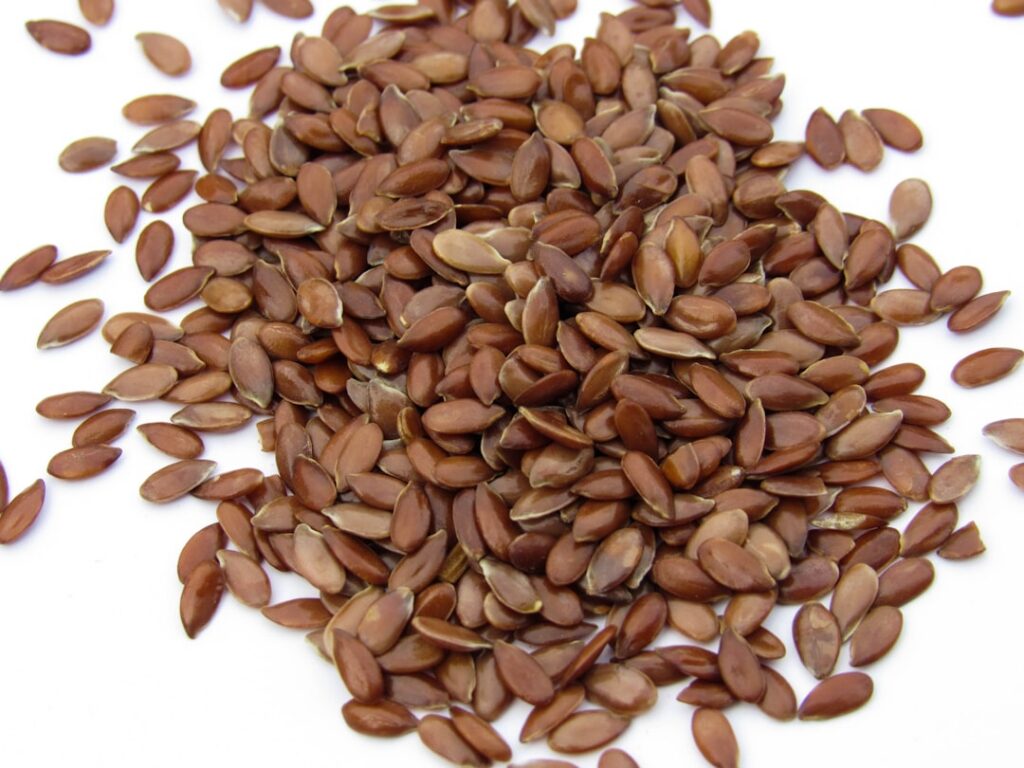 A pile of whole golden flax seeds on a wooden surface with a small measuring spoon nearby