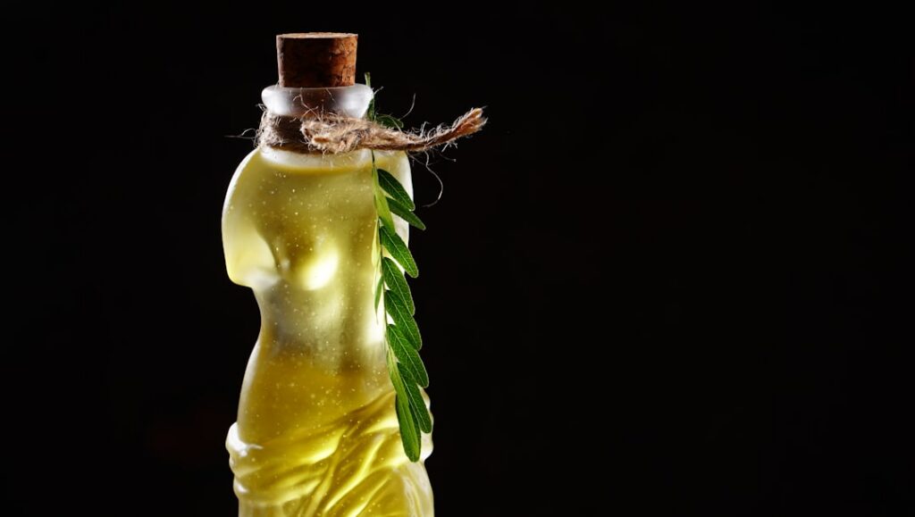 A small clear glass bottle filled with yellowish-orange infused hair oil with blurred background elements suggesting nat