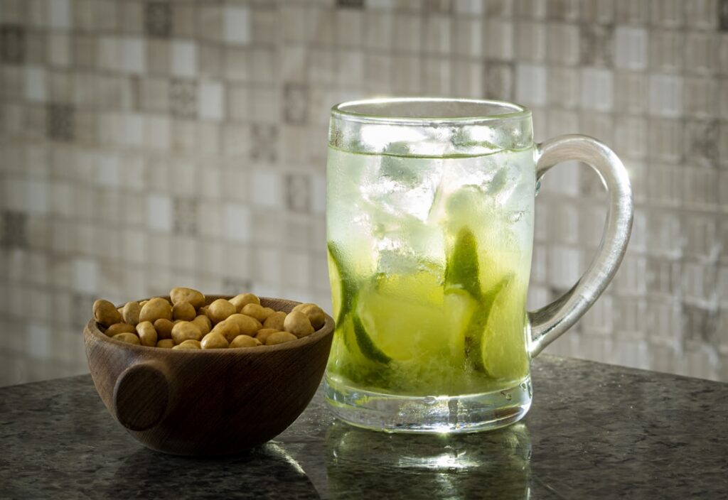 A clear glass of amber-colored fenugreek water next to a small bowl of soaked fenugreek seeds with a few raw peanuts sca