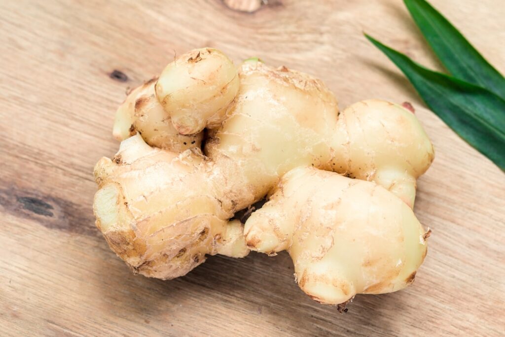 Close-up of a hand slicing fresh ginger root on a wooden cutting board with other ingredients like garlic and turmeric i