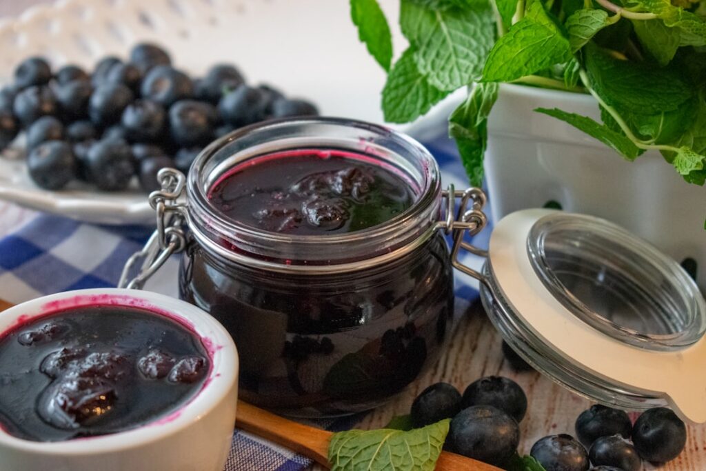 A clear glass jar filled with vibrant purple grape hyacinth syrup with a metallic spoon dripping the syrup placed on a r