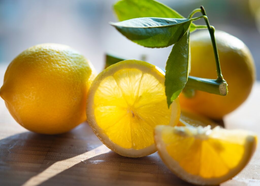 A clear spray bottle filled with a yellowish lemon seed infusion next to a fresh lemon and a pile of lemon seeds