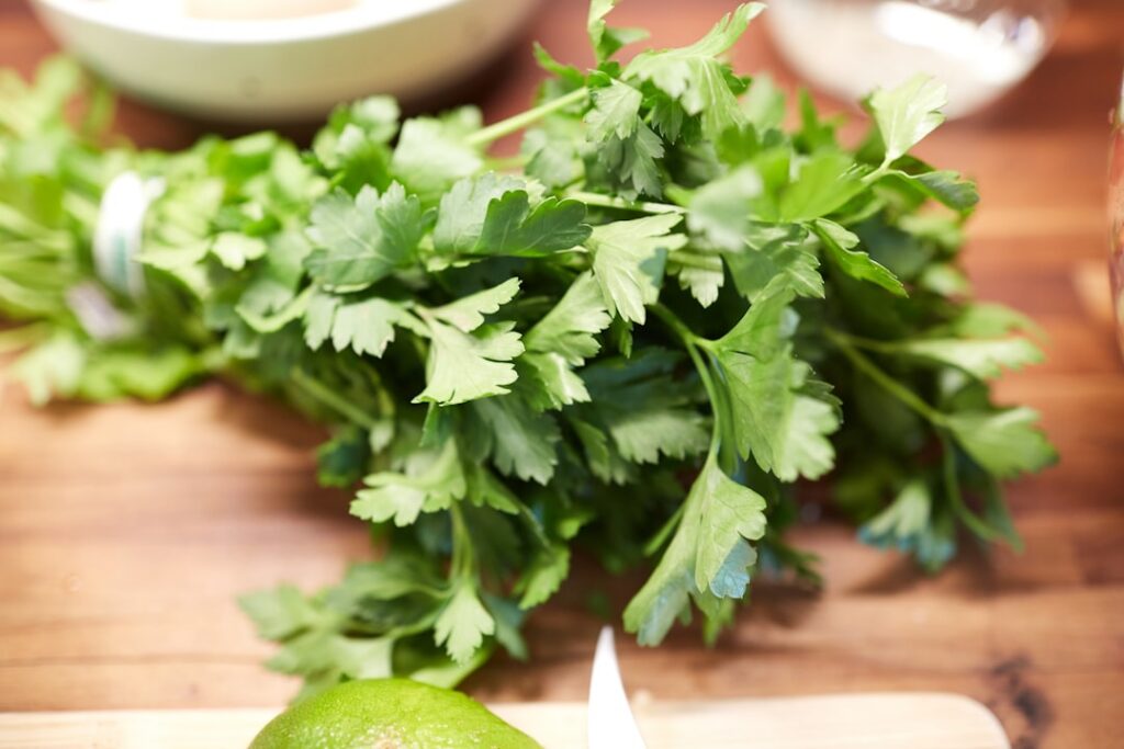 A close-up shot of a handful of fresh curly parsley and celery stalks on a rustic wooden table with a sliced lime ready 