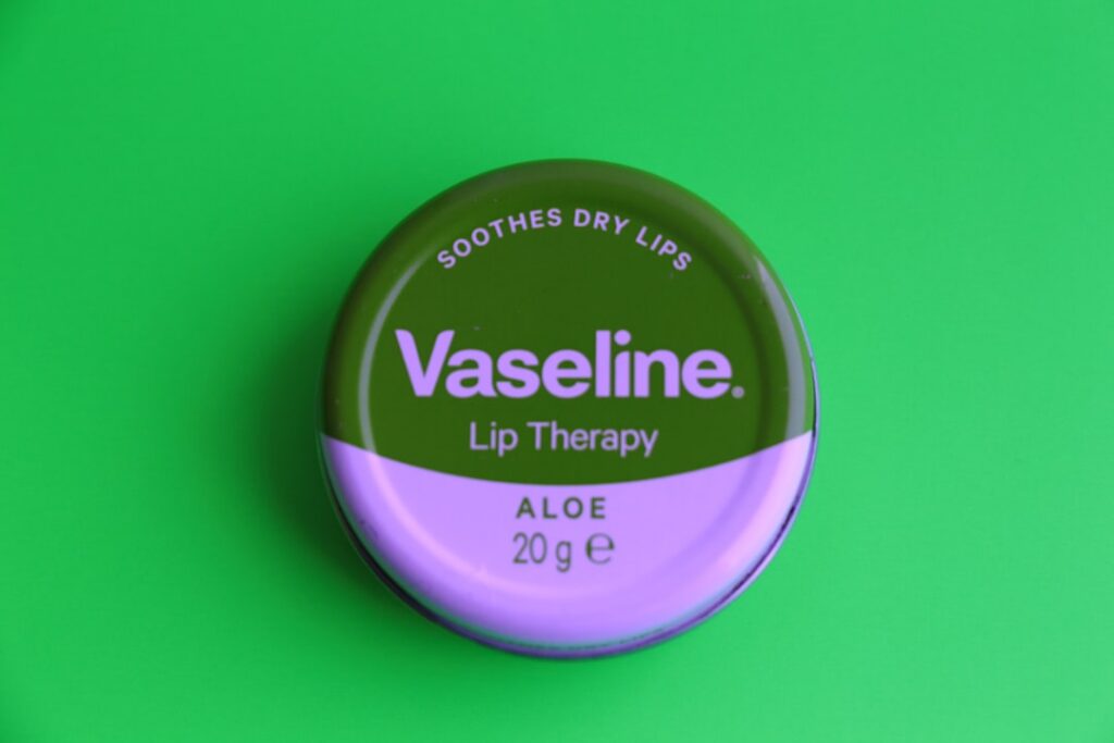 A close-up of a small glass jar containing a creamy Vaseline and aloe vera mixture with fresh aloe vera leaves next to i