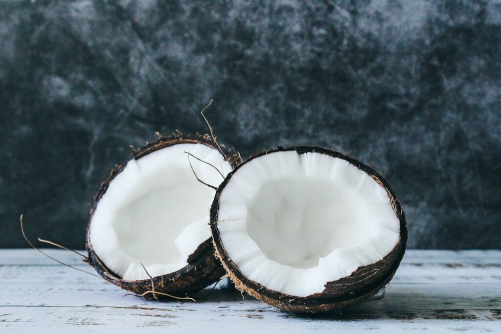 Fresh coconut halves and a bowl of creamy coconut milk surrounded by various healthy ingredients like root crops vegetab