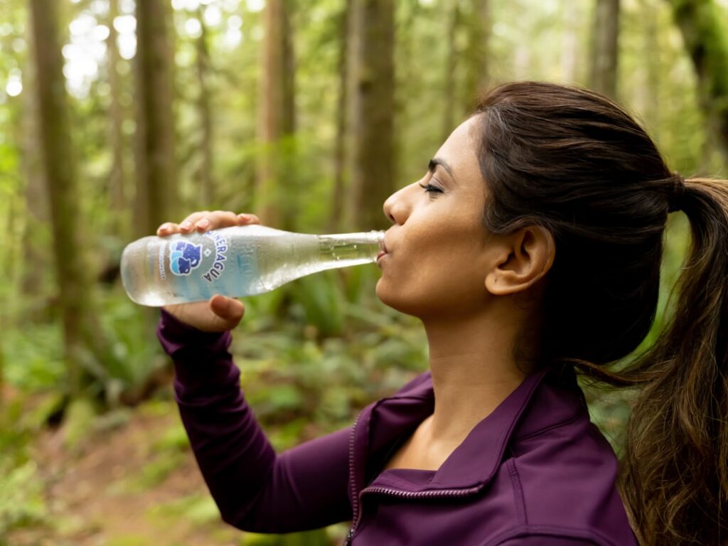 A person happily drinking water from a clear glass bottle in a bright natural setting symbolizing health and hydration