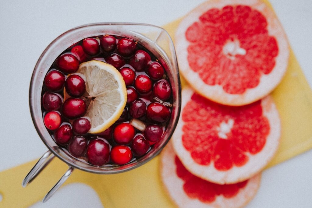 A glass of water with fresh lemon slices and a few cranberries next to a bowl of fresh lemons and cranberries