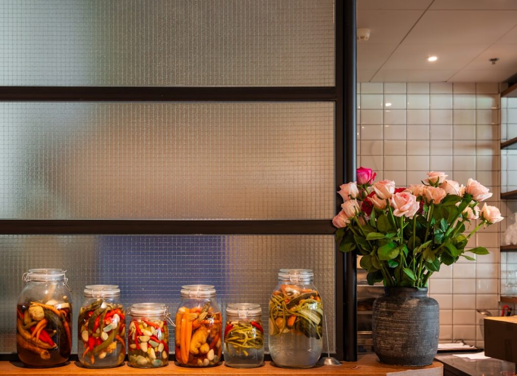 A variety of colorful pickled vegetables in small clear glass jars on a rustic wooden table with fresh herbs and spices 