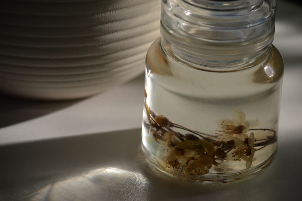 A clear glass jar filled with baby oil and dried rose petals chamomile flowers and orange peels sitting in natural sunli