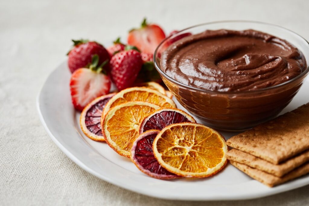 A beautiful close-up of a rich dark chocolate vegan black bean brownie dip in a bowl surrounded by fresh fruit slices li