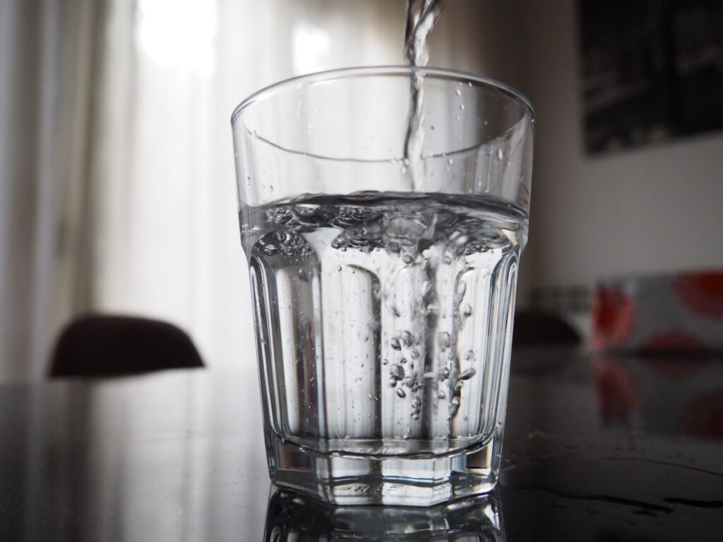 A hand pouring infused water from a glass pitcher into a clear drinking glass with fresh fruit and herb slices visible i