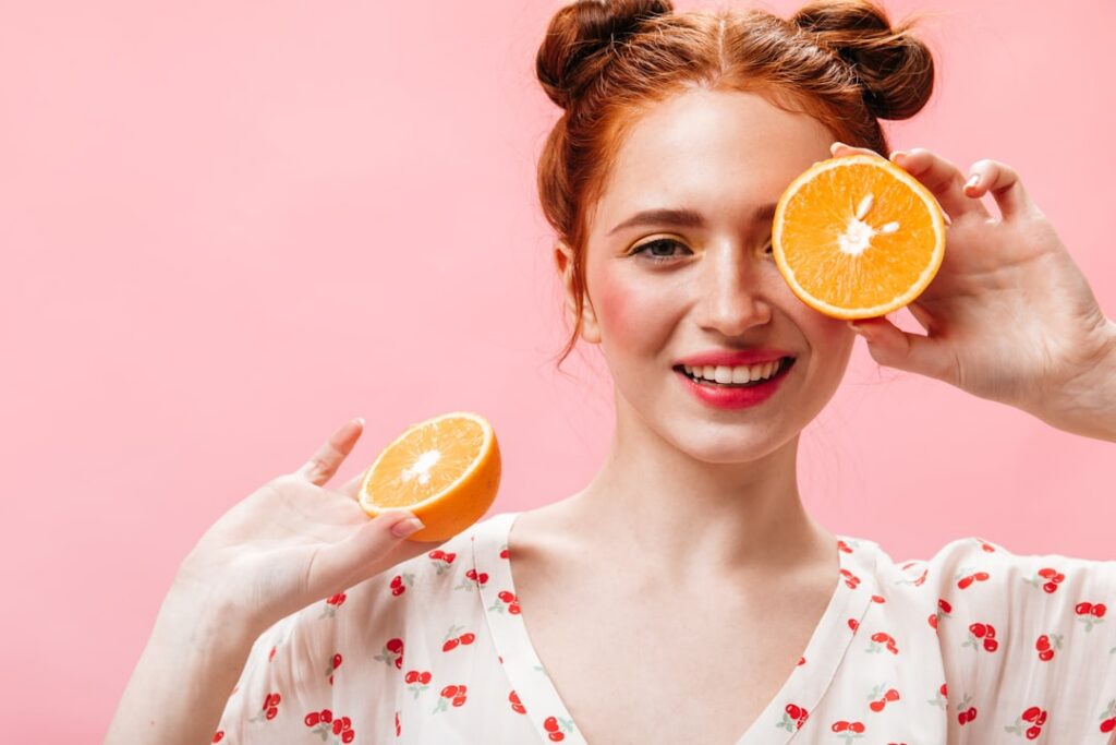 A smiling woman holding a half orange and a Vitamin C serum bottle representing healthy glowing skin after consistent da