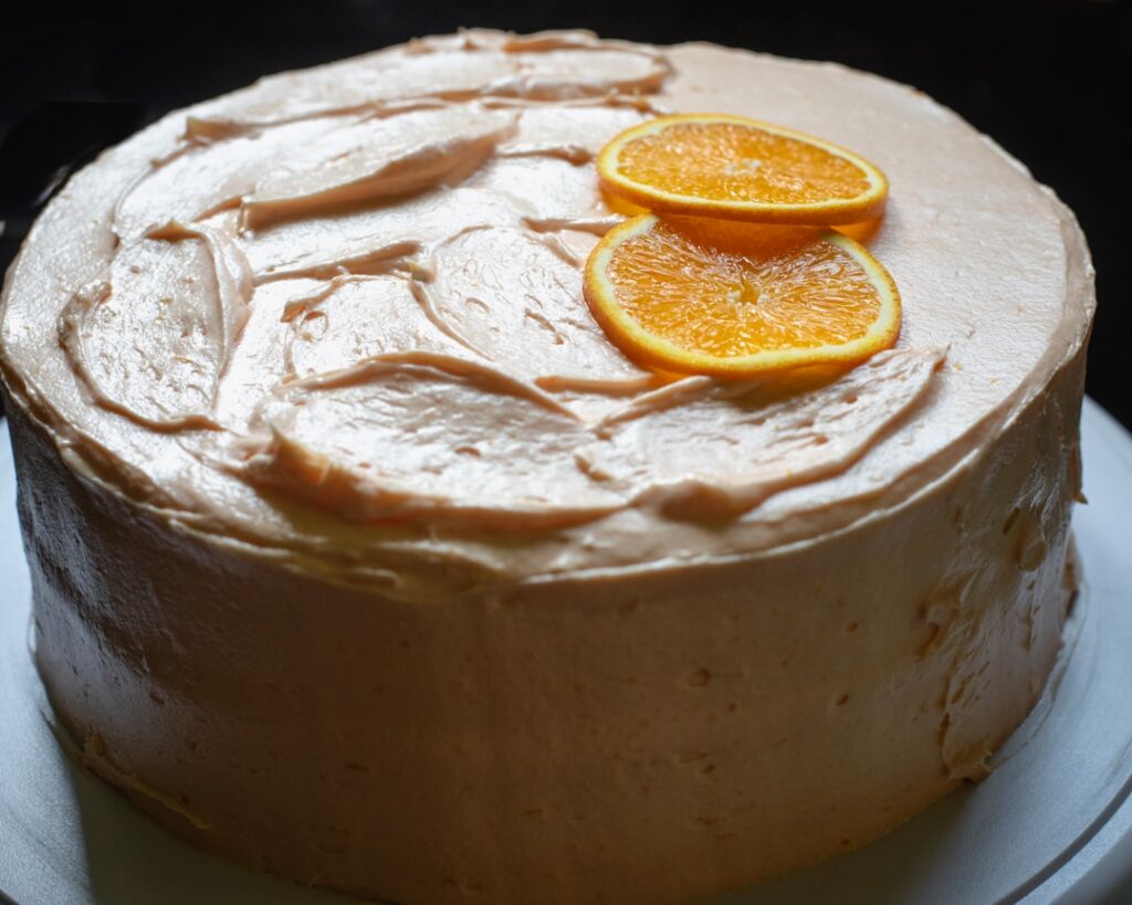 A beautifully frosted orange cake with cream cheese frosting garnished with fresh orange slices and zest on a cake stand