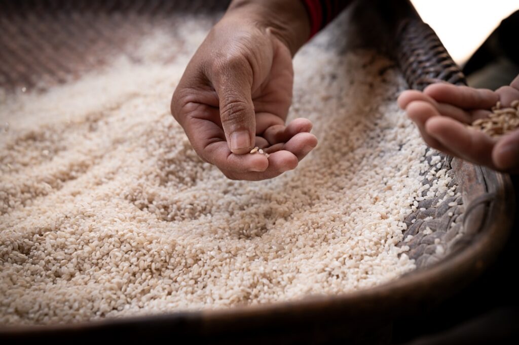 A close-up of a glass jar filled with uncooked rice and water being stirred by a hand illustrating the soaking method fo
