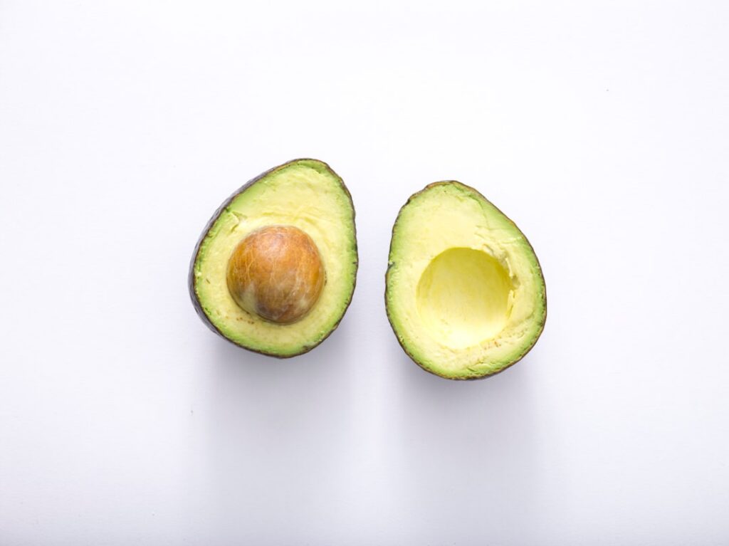 A vibrant green avocado half and a small bowl of olive oil on a wooden surface ready for making a hair mask