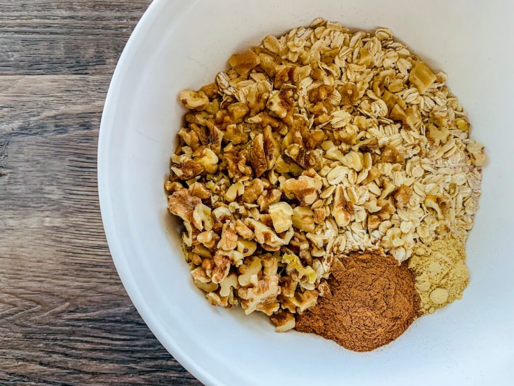 Various dry oatmeal toppings like nuts seeds and dried fruits in small bowls alongside a container of rolled oats and me