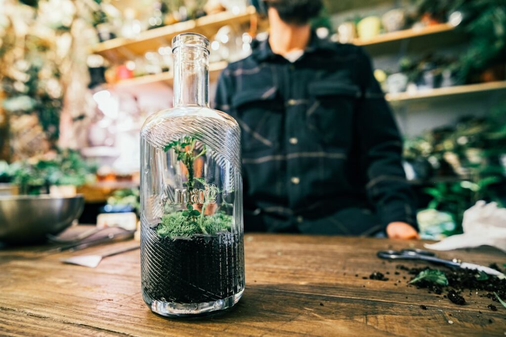 A pair of hands using long tweezers to carefully plant a small fern into the soil layer of a partially built terrarium
