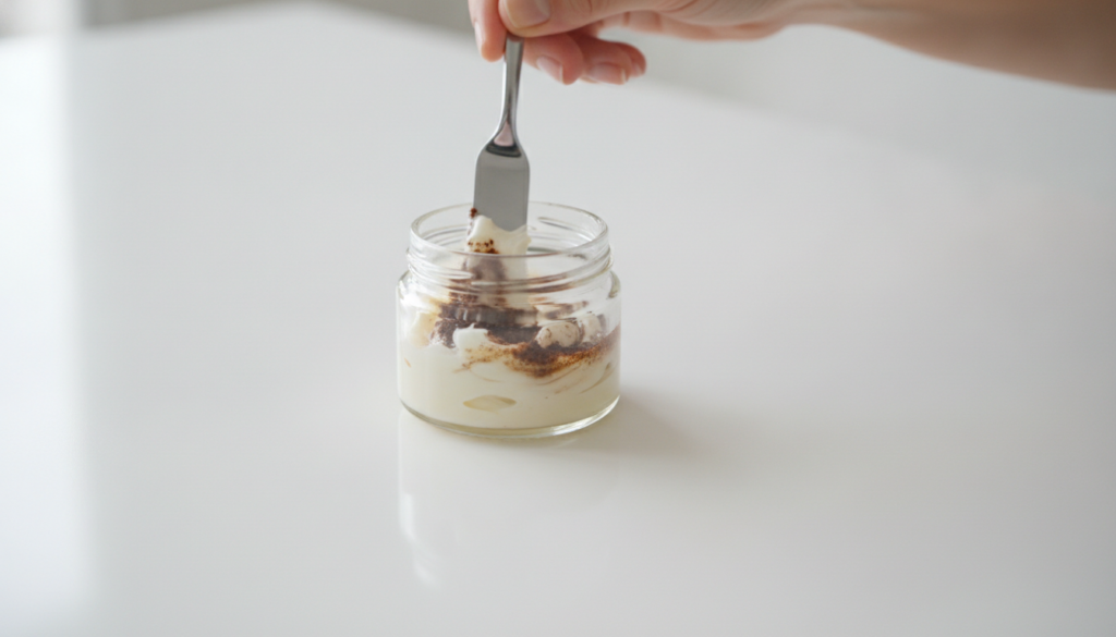 A close-up of a small jar of Vaseline with ground coffee being mixed into it by a finger or small spatula against a soft