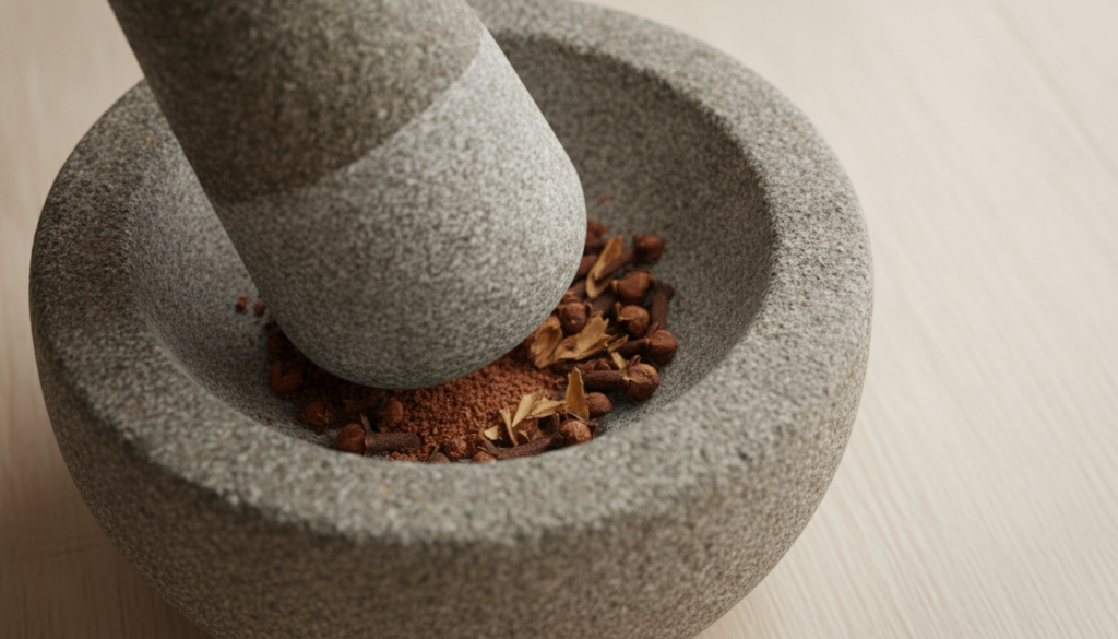 A close-up of whole cloves being gently crushed in a small mortar and pestle showing some broken pieces of cloves on a c