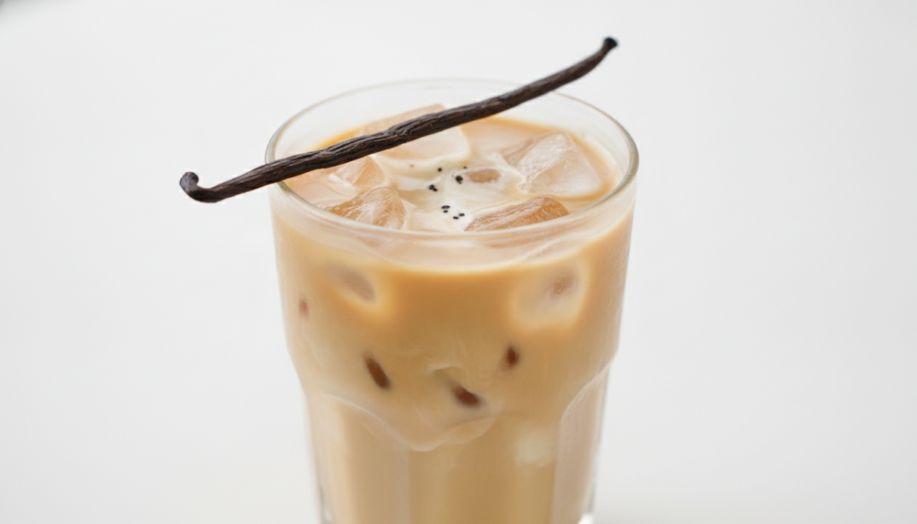 A tall glass of French vanilla iced coffee with a vanilla bean pod as garnish The coffee is light brown with ice cubes a