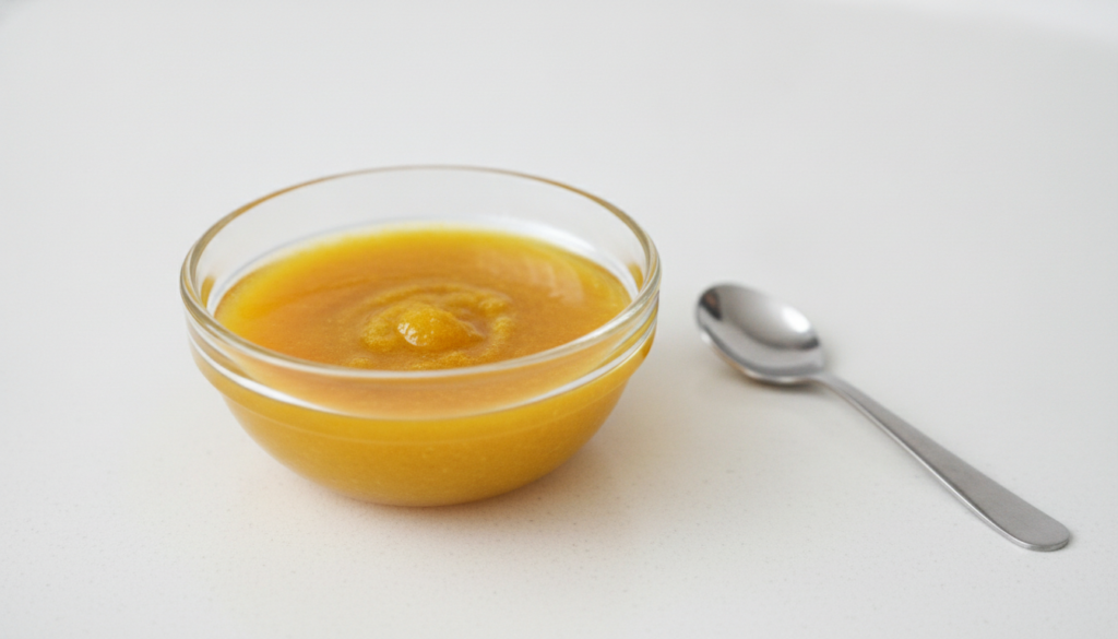 A close-up of a small glass bowl with a golden-yellow mixture of baby oil and turmeric with a small mixing spoon or brus