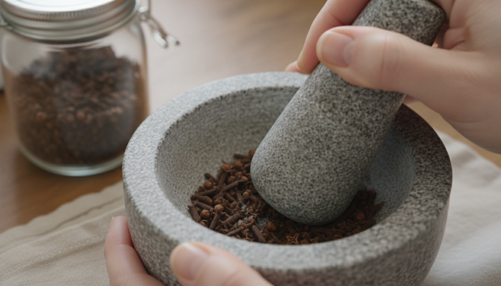 Hands gently crushing whole cloves with a mortar and pestle next to a glass jar ready for oil infusion