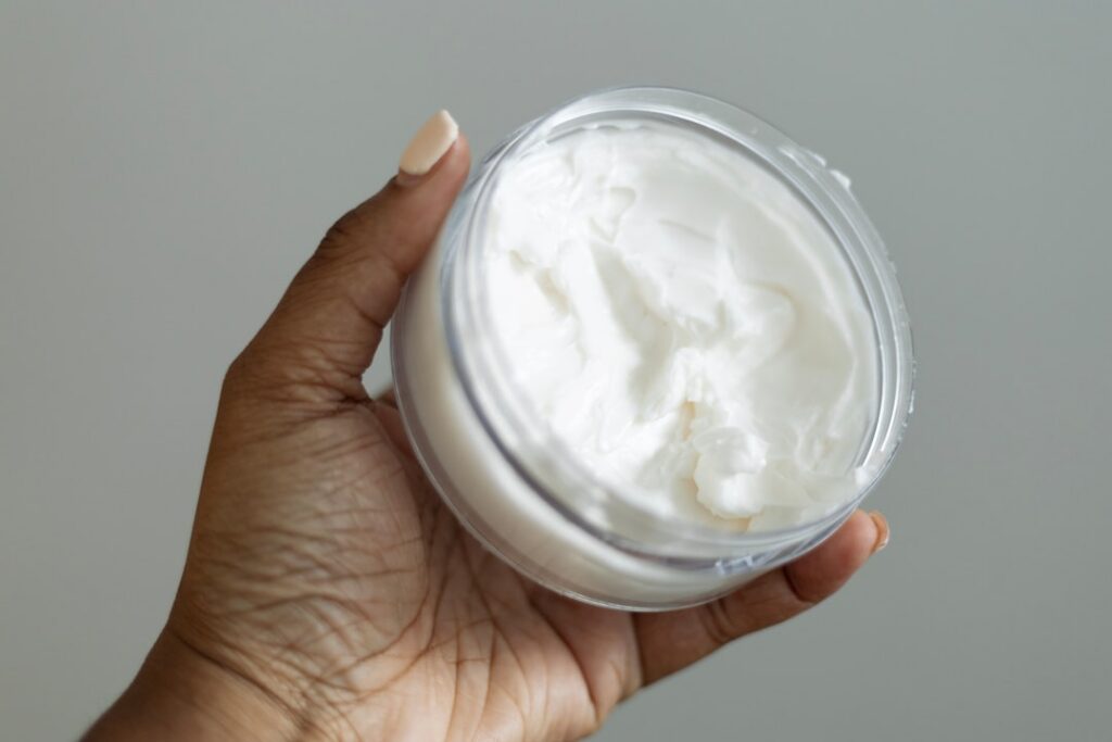 A close-up of a small elegant glass jar filled with a homemade cream with Vitamin E capsules and a tube of Vaseline visi