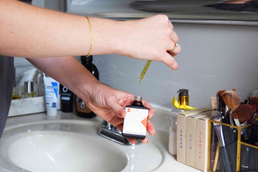 A person s hand holding a glass spray bottle filled with a clear liquid labeled Bathroom Mist and spritzing it into the