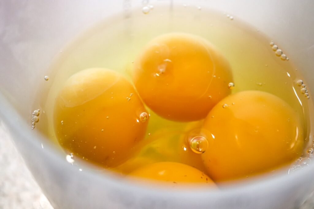 Close-up shot of a small bowl with egg yolk and baby oil a whisk and a small jar of honey on a clean light surface The i