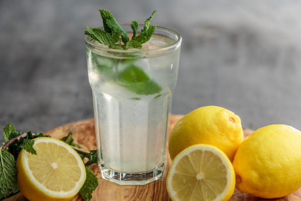 A glass of vibrant yellow turmeric water with a slice of lemon and a sprig of mint on a light wooden table with a bowl o