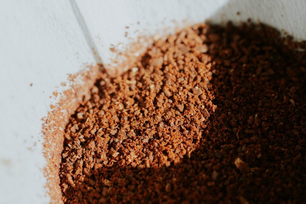 A close-up shot of used coffee grounds being sprinkled around the base of a healthy green plant in a garden bed