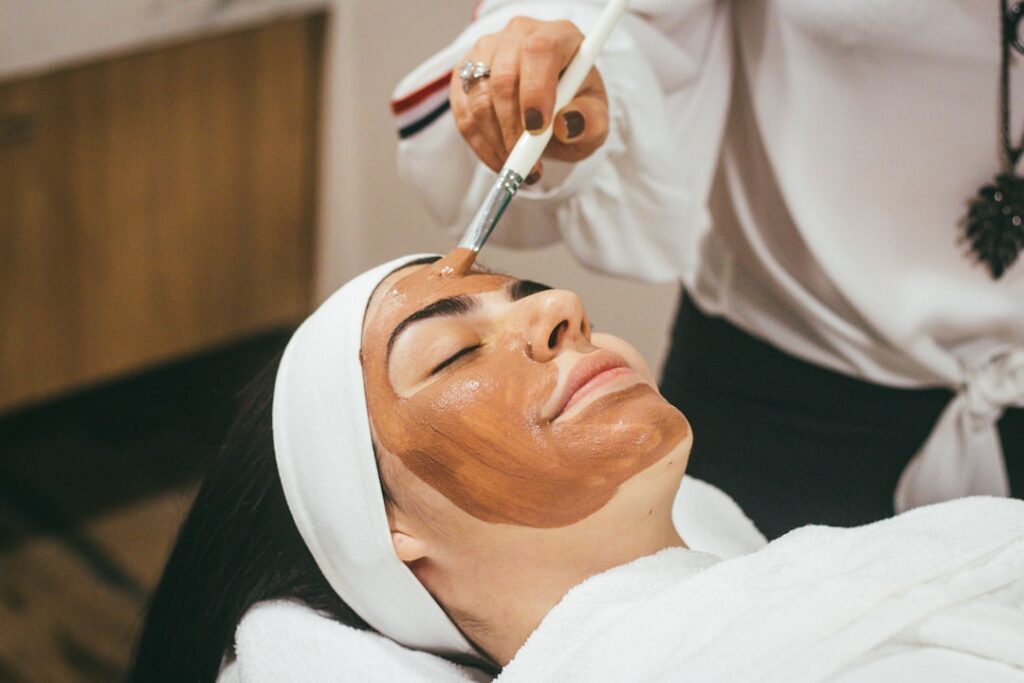 A woman gently applying a smooth light brown herbal paste to her clean face with a soft smile in a brightly lit bathroom