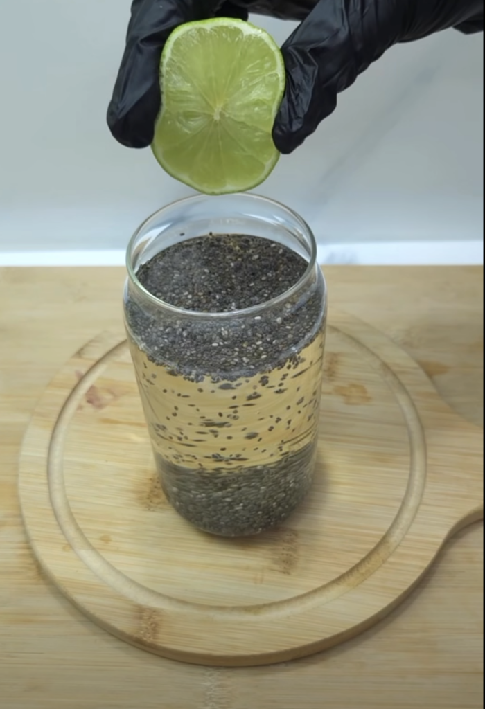 A hand wearing a black glove squeezing a fresh lime half over a glass jar containing a light-colored liquid with chia se