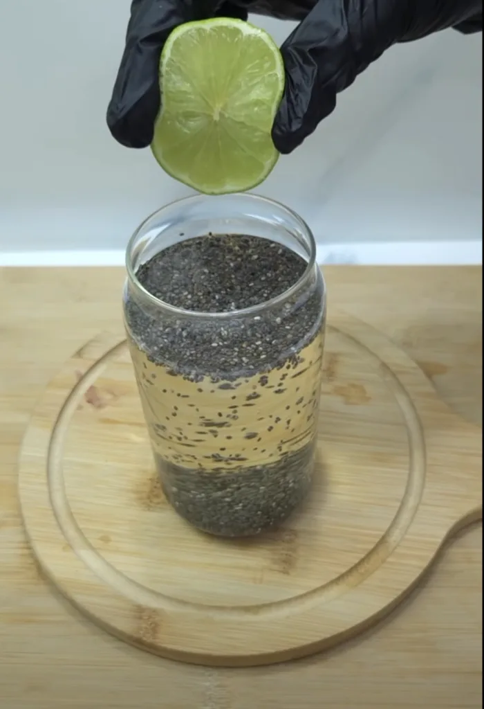 A hand wearing a black glove squeezing a fresh lime half over a glass jar containing a light-colored liquid with chia se