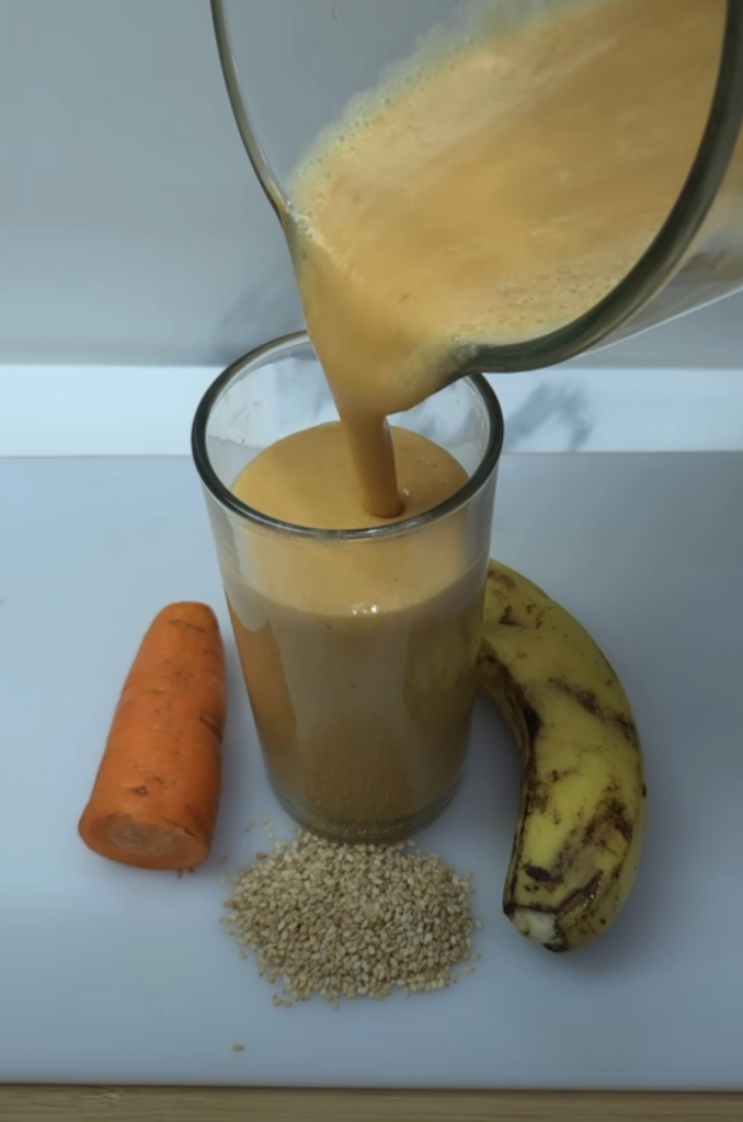 A creamy orange smoothie being poured from a blender into a clear glass with a whole carrot a peeled banana and a small