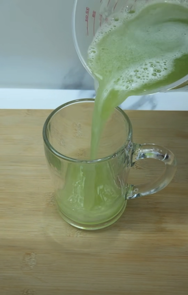 A vibrant green liquid is being poured from a clear measuring cup into a clear glass mug creating a frothy top layer The