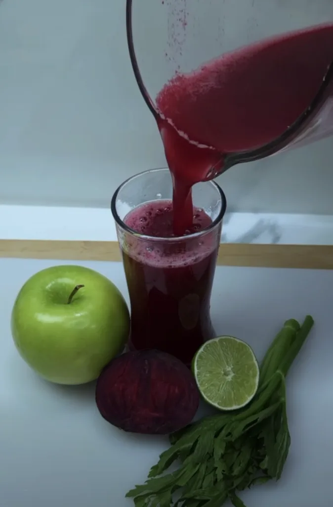 A vibrant rich red juice being poured from a clear pitcher into a tall glass surrounded by fresh ingredients like a gree