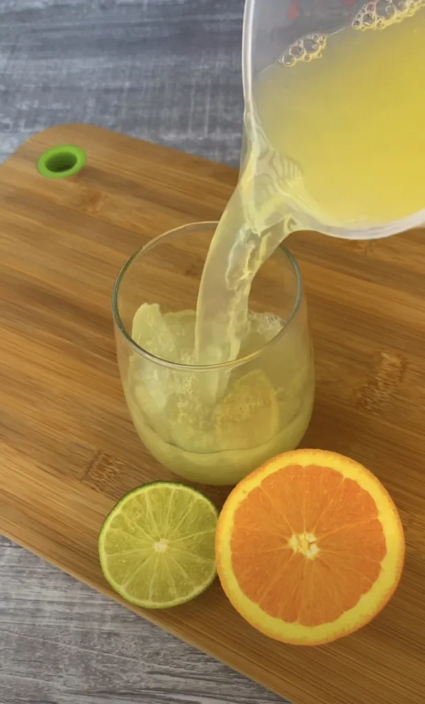 A vibrant light yellow citrus drink being poured from a clear pitcher into a glass already partially filled On a wooden 
