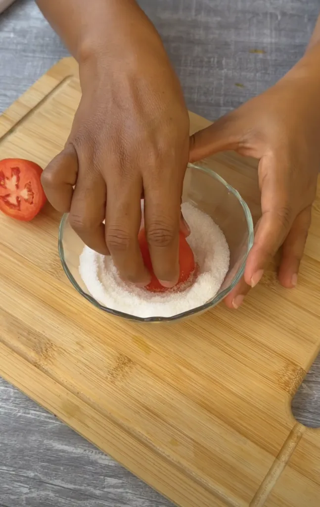 A pair of hands gently pressing a halved red tomato into a clear glass bowl filled with white sugar set on a light brown