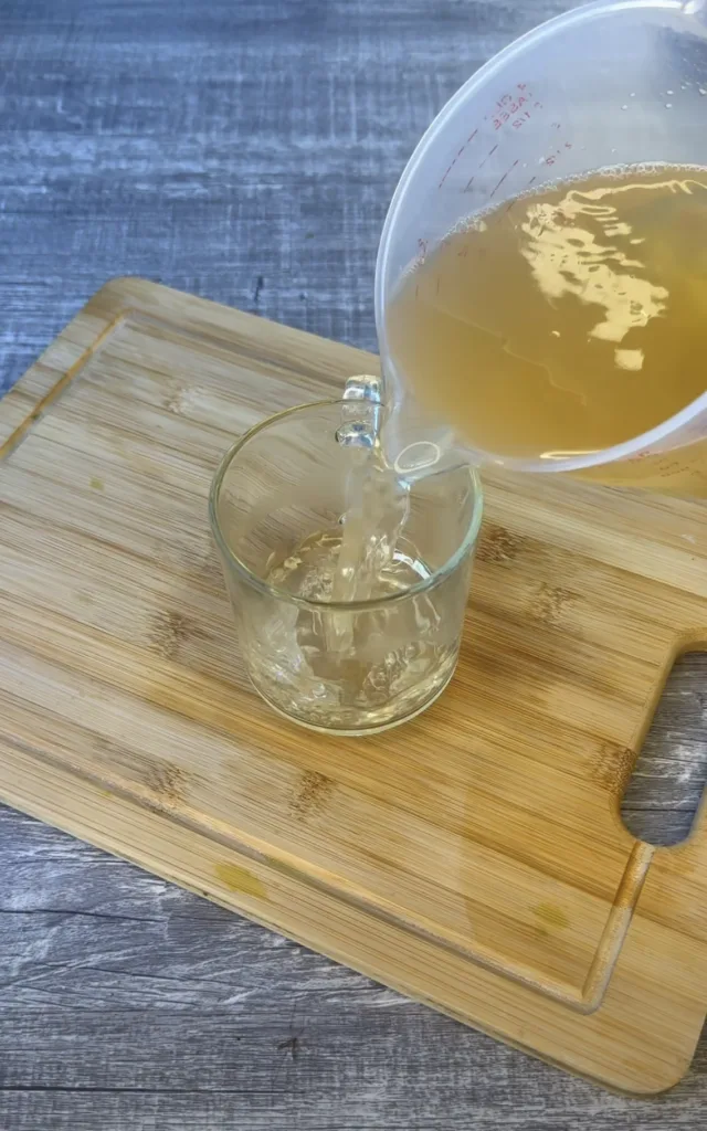 Overhead shot of amber liquid being poured from a measuring cup into a glass on a wooden cutting board