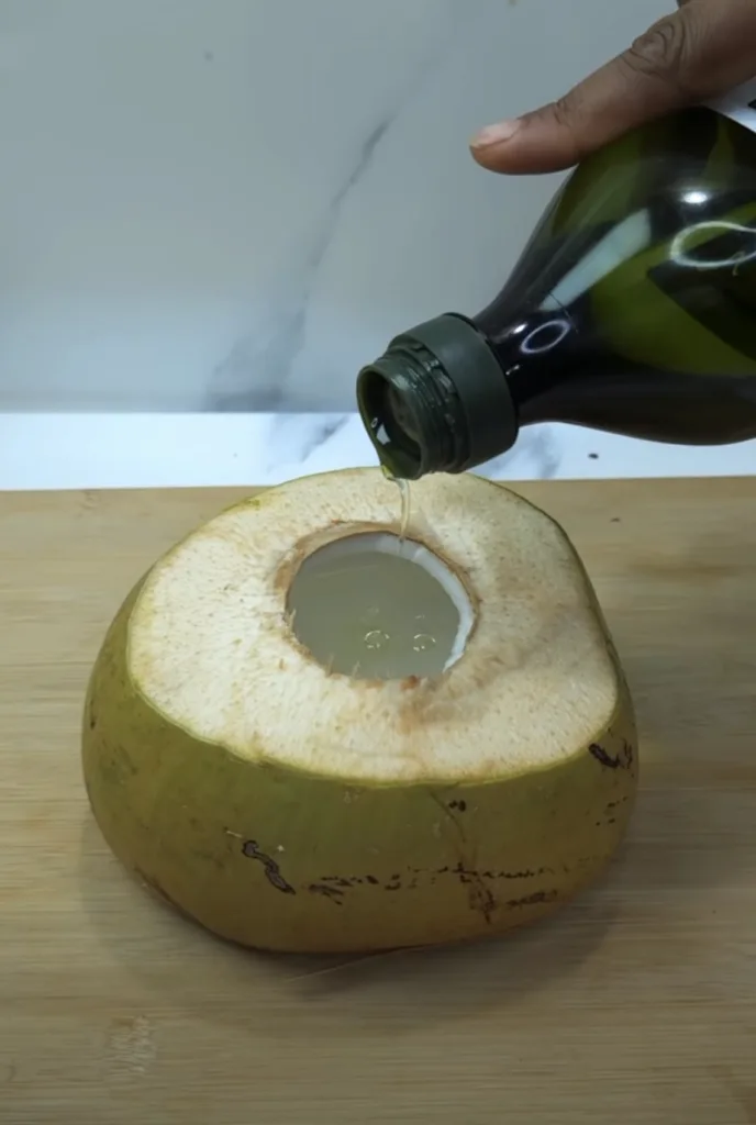 A hand pouring a rich dark liquid from a green bottle into a hollowed-out green coconut on a wooden cutting board