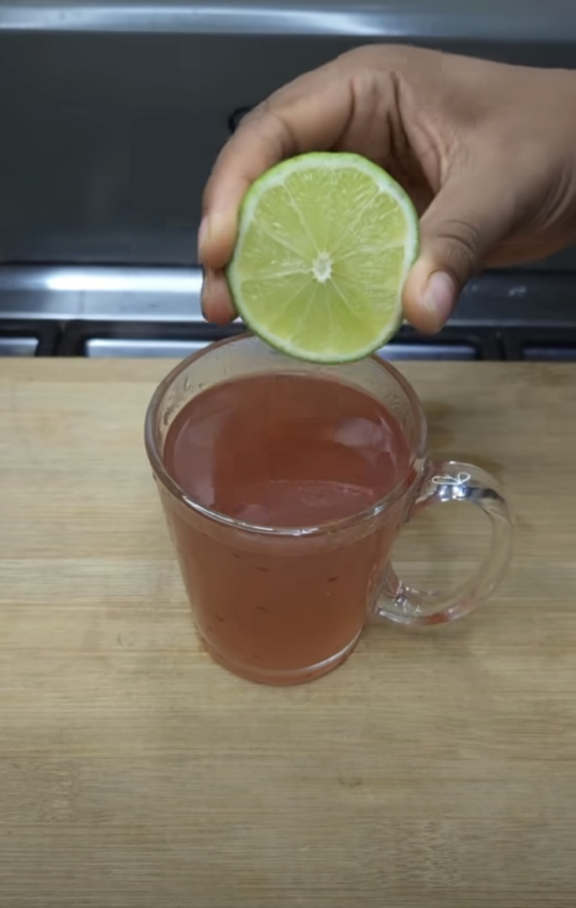 A hand is squeezing a half-sliced lime into a clear glass mug containing a reddish-pink beverage with a blurred kitchen 