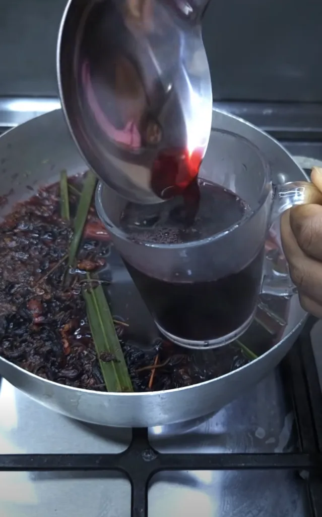 A close-up shot of a dark red herbal beverage being poured from a metal ladle into a clear glass mug In the background a