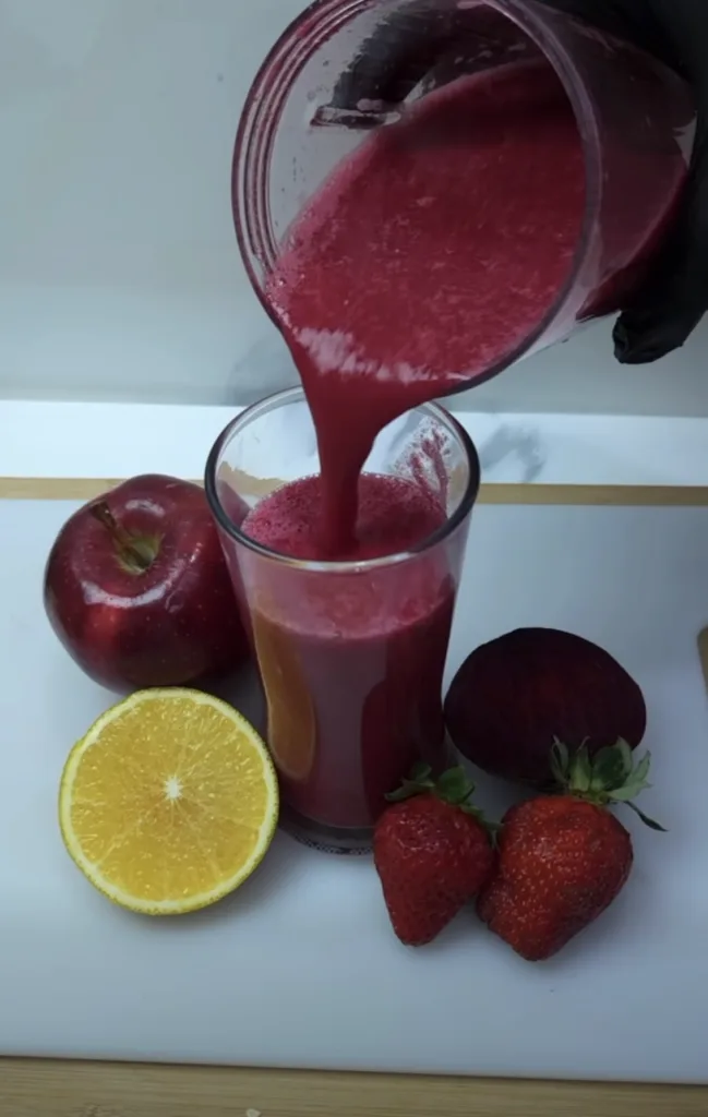 A vivid red fruit and vegetable juice being poured from a blender into a clear glass with a red apple half an orange a w