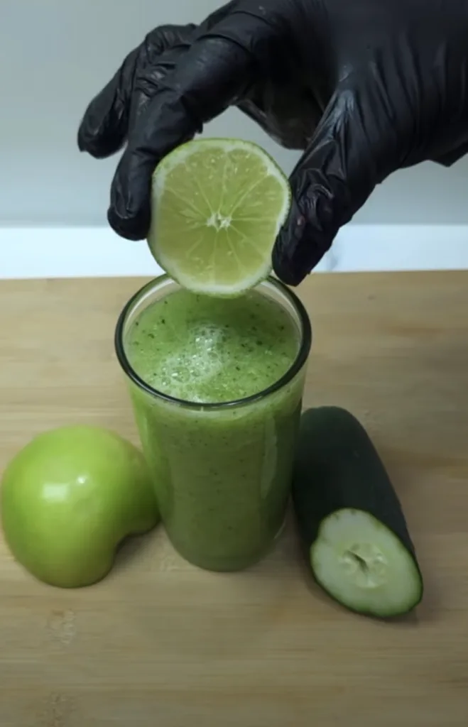 A hand in a black glove holding a fresh lime half directly over a tall glass of vibrant green smoothie with a foamy laye