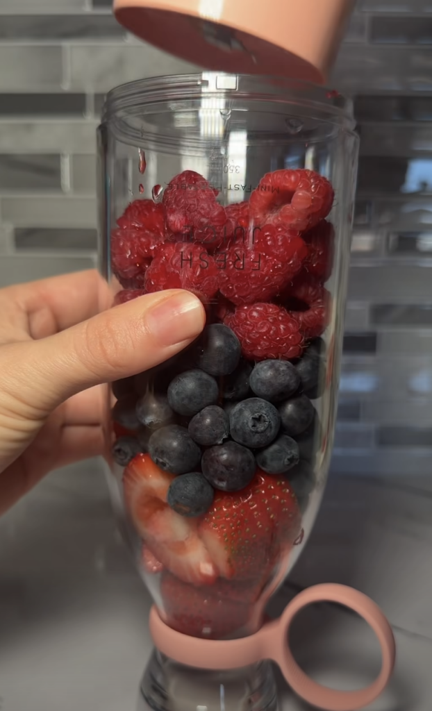 A clear blender bottle filled with fresh raspberries blueberries and sliced strawberries held by a hand A peach-colored 