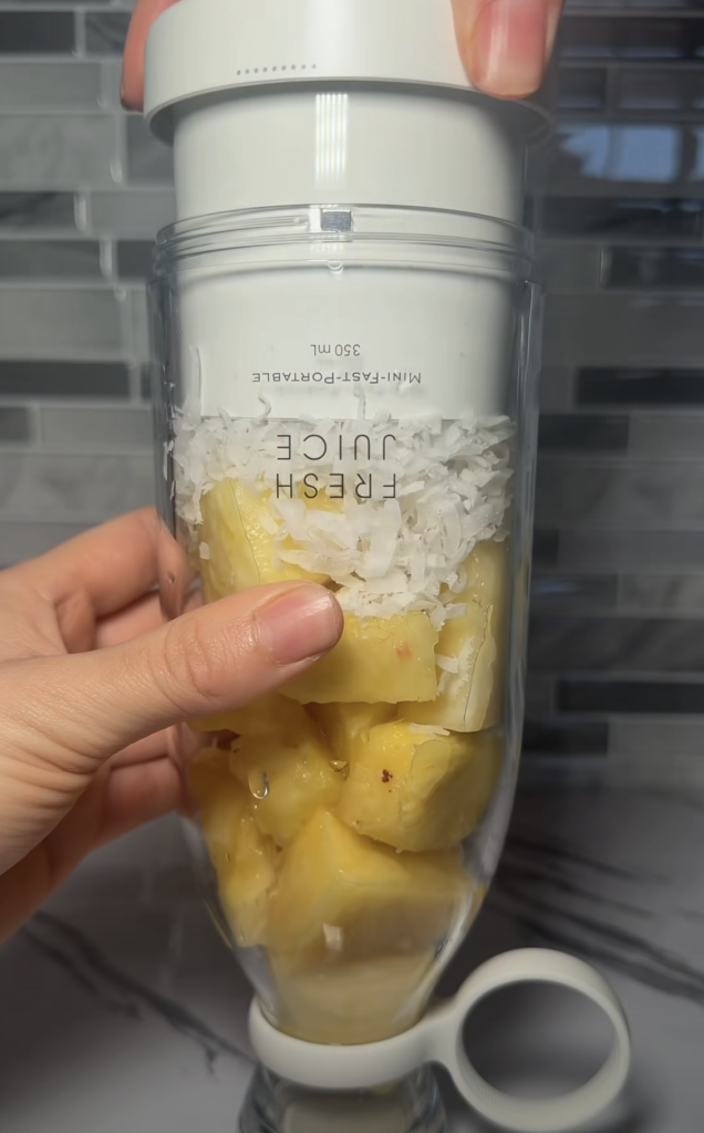 A clear portable blender filled with chunks of fresh pineapple and a layer of shredded coconut on top held by a hand
